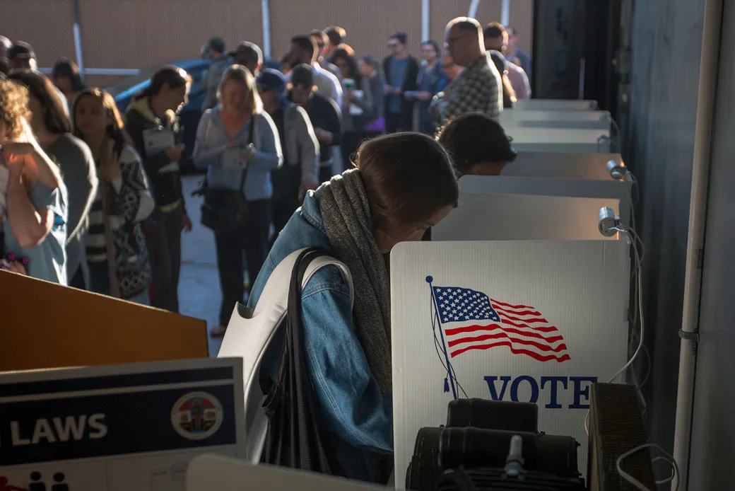 Voters standing in line at polling booths on election day, with one woman in a blue jacket and gray scarf focused at her voting station; booths feature 'VOTE' signs, American flags, and privacy curtains in an indoor polling place.